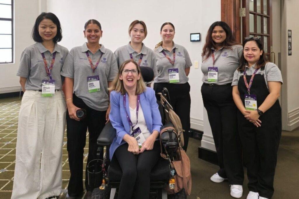 Six women pose indoors, five standing and wearing matching grey polo shirts with name badges, and one woman seated in a wheelchair, smiling and wearing a light blue blazer. They are in a conference setting.