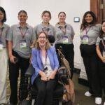 Six women pose indoors, five standing and wearing matching grey polo shirts with name badges, and one woman seated in a wheelchair, smiling and wearing a light blue blazer. They are in a conference setting.