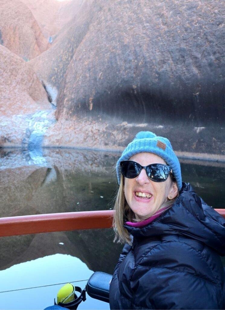 A woman in sunglasses and a blue beanie smiles at the camera while sitting in a power chair near a body of water, with rocky cliffs and a small waterfall in the background.