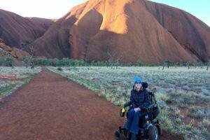 A woman in a power wheelchair smiles at the camera on a dirt path with Uluru, a large red sandstone monolith, and open grassy landscape in the background under a clear sky.
