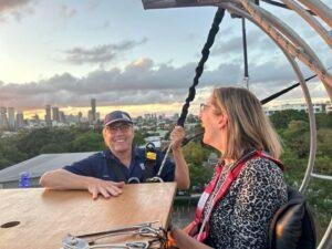 Two people wearing harnesses smile and talk at a high outdoor dining table, with a city skyline, trees, and a sunset in the background. One person holds a safety strap; diners are elevated above the ground.