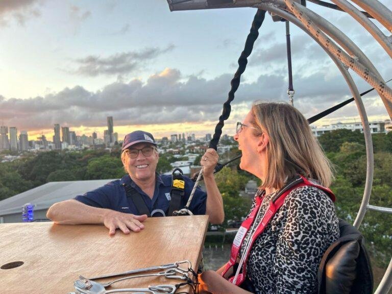 Two people wearing harnesses smile and talk at a high outdoor dining table, with a city skyline, trees, and a sunset in the background. One person holds a safety strap; diners are elevated above the ground.