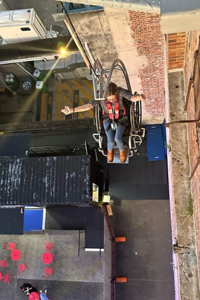 A women sitting in a wheelchair wearing a harness is suspended high above the ground by a mechanical ride on the side of a brick building, giving a thumbs-up. Below are shipping containers, tables with red chairs, and urban surroundings.