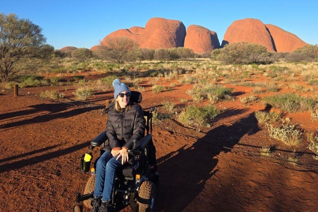 A smiling woman wearing sunglasses, a beanie, and a black jacket sits in a power wheelchair on red soil, with grass, trees and large red rock formations (Kata Tjuṯa) in the background.