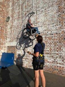 A smiling woman in a wheelchair is being lifted up a brick wall using an adaptive pulley system, assisted by a person wearing a safety harness. Sunlight casts their shadows on the wall.