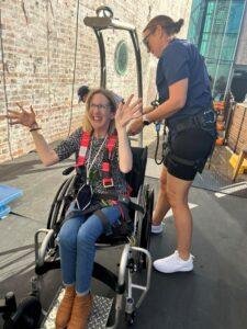 A smiling woman in a wheelchair, wearing a harness and raising her hands in excitement, is assisted by another woman in a harness. They are outdoors near a brick wall.