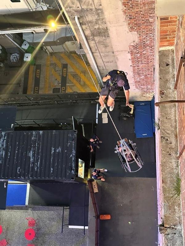A person in uniform rappels down a brick wall with a rope, while two people on the ground look up and guide. Various objects, including mats and a metal structure, are visible below in an urban setting.