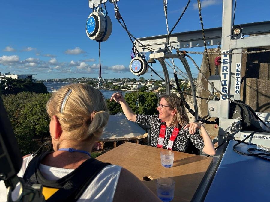 Two women wearing safety harnesses sit at a raised outdoor table with drinks, suspended high above the ground, enjoying the view on a sunny day. Buildings, trees, and blue sky are in the background.
