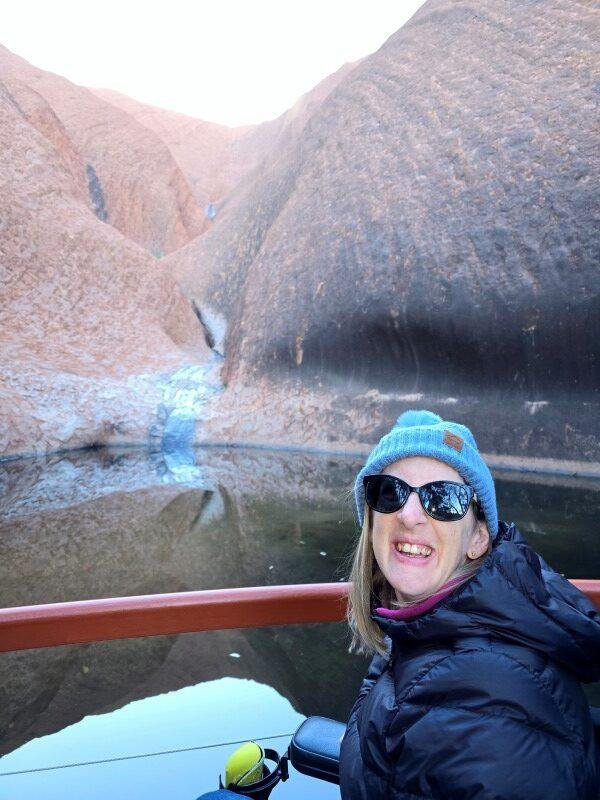 A woman wearing sunglasses, a blue beanie, and a black jacket smiling near a body of water with rocky hills in the background.