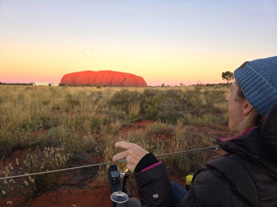 A womanwearing a blue beanie and black jacket sits outdoors, holding a glass of white wine, facing Uluru at sunset with a clear sky above and Australian outback vegetation in the foreground.