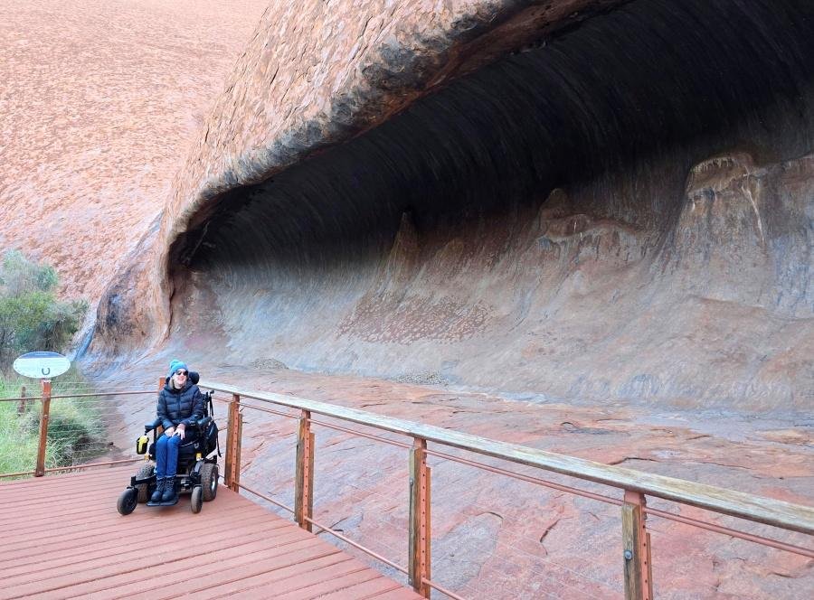 A woman in a power wheelchair sits on a wooden accessible pathway beside a large, curved rock formation at Uluru, Australia. The scene includes a railing and natural rock textures in the background.