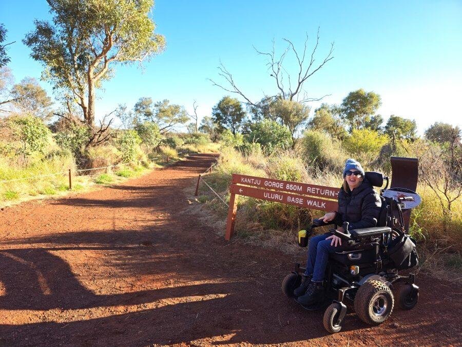 A woman in a power wheelchair smiles near a brown sign on a dirt path, surrounded by green trees and bushes under a clear blue sky. The sign points to Katju Gorge and Uluru Base Walk.