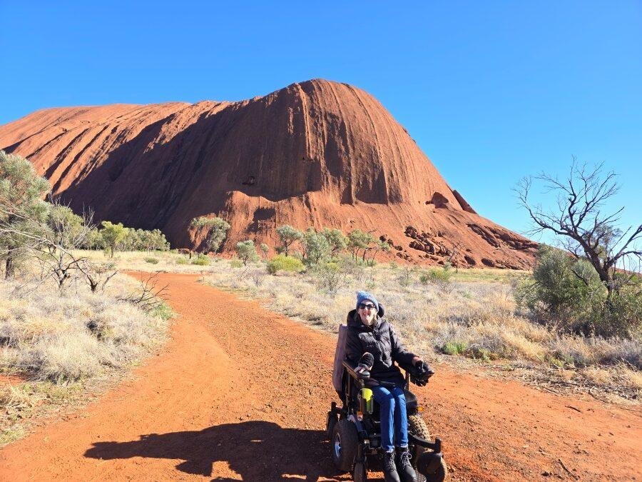 A woman in a power wheelchair is on a red dirt path with Uluru, a large red rock formation, in the background under a clear blue sky, surrounded by dry grass and scattered trees.