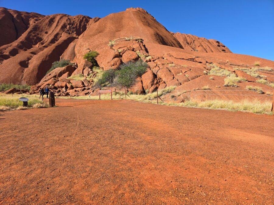 A large red sandstone rock formation rises under a clear blue sky, with a dirt path and sparse vegetation in the foreground. A few people are walking near the base of the rock.