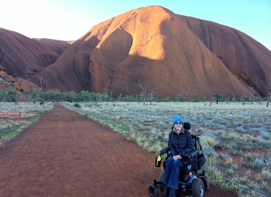 A woman in a power wheelchair sits on a red dirt path with sparse grass, smiling and wearing a blue bennie, black jacket and sunglasses. Behind her is the large, sunlit red rock formation of Uluru.