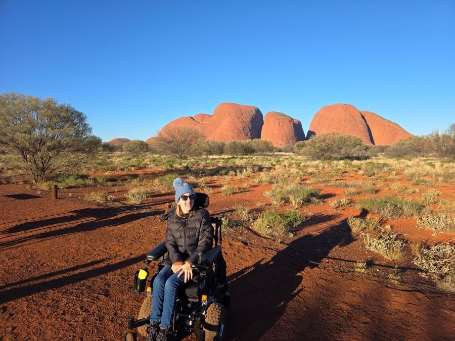 A women in a power wheelchair sits on a red dirt path in front of large, rounded rock formations under a clear blue sky, surrounded by sparse desert vegetation.
