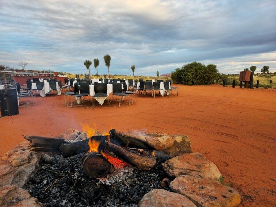 A stone firepit with burning logs in the foreground, surrounded by red dirt. In the background, tables and chairs with white tablecloths are set up outdoors under a cloudy sky, with sparse trees and shrubs nearby.