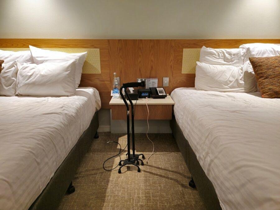 Two beds with white bedding are separated by a small nightstand holding a phone, water bottle, and lamp. A black walker is positioned between the beds on a beige carpet.