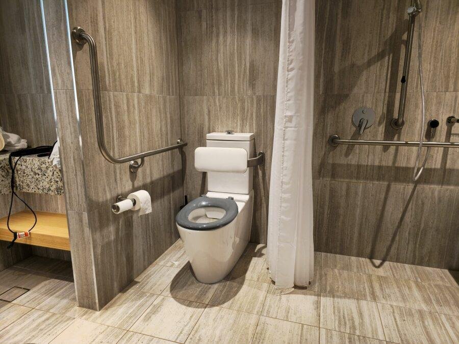 An accessible bathroom with a toilet featuring grab bars, a backrest, a roll of toilet paper, a shower area with a handheld showerhead, and a curtain. The floor and walls are tiled.
