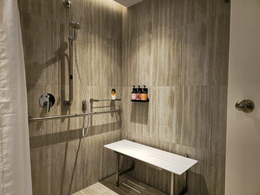 Accessible shower with a handheld showerhead, horizontal grab bar, wall-mounted soap dispensers, and a white bench on a tiled wall.