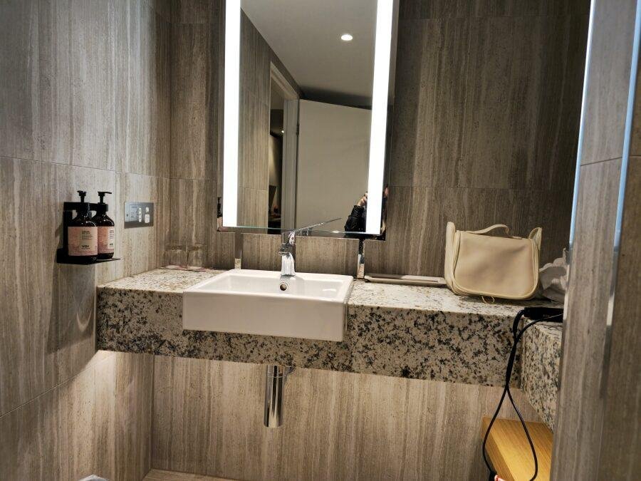 A modern bathroom with gray tiled walls, a rectangular illuminated mirror, a square sink on a granite countertop, soap dispensers, a beige bag, a hairdryer, and a glass placed near the faucet.