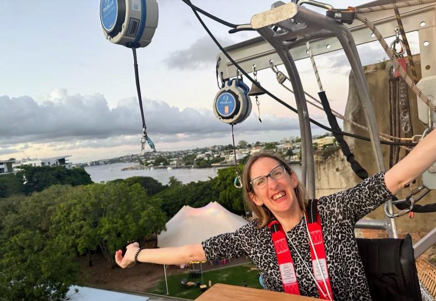 A smiling woman sitting in a wheelchair and arms outstretched sits in a harness on an outdoor platform, with trees, a river, and a cityscape in the background under a cloudy sky.