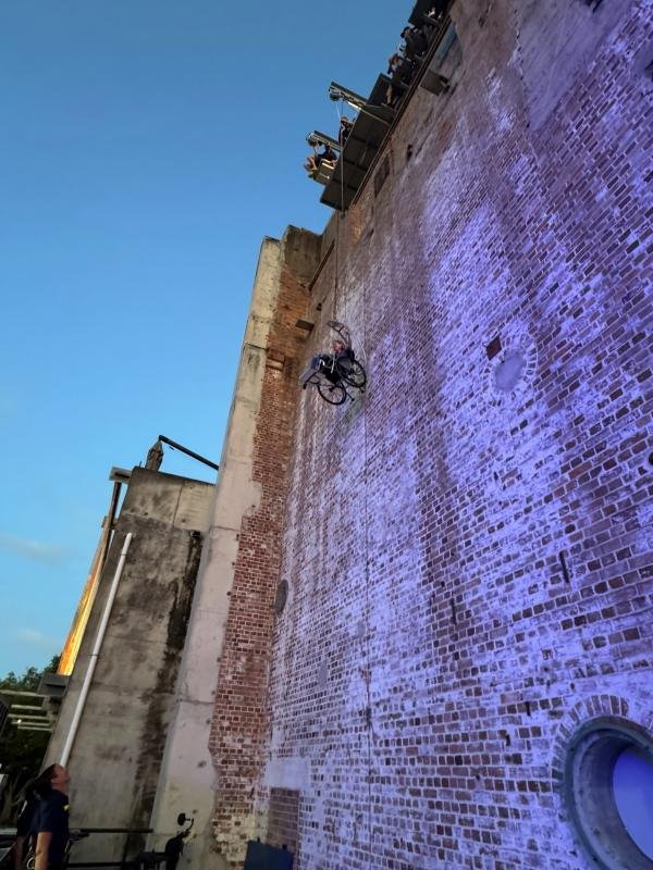 A woman in a wheelchair rides vertically down a tall brick wall, with people watching from above and below against a clear evening sky.