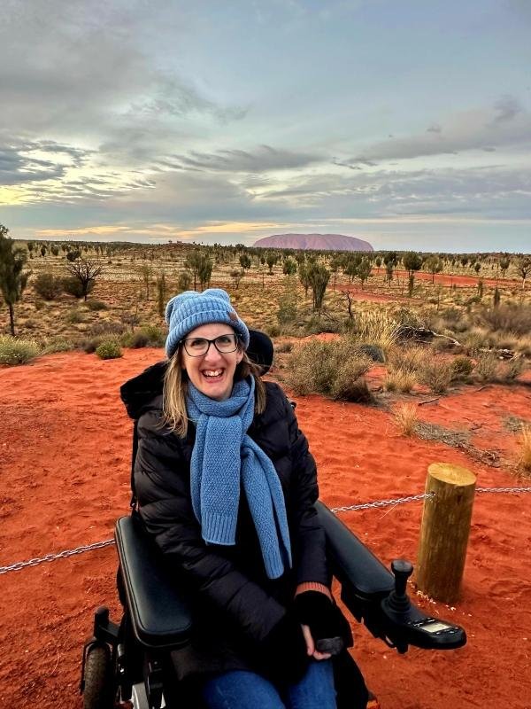 A woman in a power wheelchair, wearing a blue bennie, scarf, and glasses. Behind her is red desert soil, sparse shrubs, and the distant Uluru rock formation under a cloudy sky.